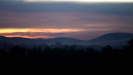 Fireworks-burst-and-sparkle-under-dusk-glow-of-sky-and-mountains-in-front-of-forest