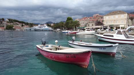 Un-Clip-De-Varios-Barcos-Amarrados-En-Un-Muelle-Local-En-Cavtat,-Croacia,-Balanceándose-Suavemente-En-Las-Cristalinas-Aguas-Azules-Del-Mediterráneo-En-Una-Tarde-Tranquila-Con-La-Ciudad-De-Fondo