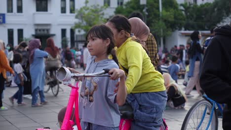 Zwei-Mädchen-Mit-Fahrrädern-Auf-Einem-Belebten-Platz-Mit-Herumlaufenden-Menschen