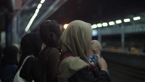 Personas-En-Una-Estación-De-Tren-Con-Poca-Luz,-Una-Sosteniendo-A-Un-Bebé,-Usando-Hijabs,-Esperando