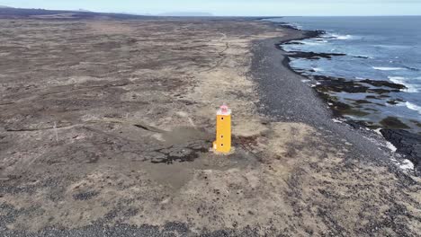 Aerial-drone-footage-pans-over-a-charming-orange-lighthouse-with-a-pink-roof,-nestled-on-a-black-sand-beach-along-Iceland's-scenic-coast