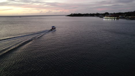 Antena-De-Un-Barco-Motorizado-Deslizándose-Sobre-El-Agua-Desde-La-Costa-Hacia-Una-Isla-Iluminada-Durante-El-Crepúsculo-En-Roatán,-Honduras