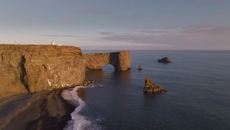 Dyrhólaey-and-black-beach-in-Iceland-with-orange-sunrise-lights,-aerial-sideways