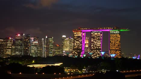 Nacht-Zeitrafferaufnahme-Im-Marina-Barrage-Rooftop-Park,-Die-Den-Beleuchteten-Supertree-Grove-In-Den-Gardens-By-The-Bay-Und-Die-Innenstadtansicht-Mit-Dem-Markanten-Marina-Bay-Sands-Vor-Schnell-Ziehenden-Wolken-Einfängt