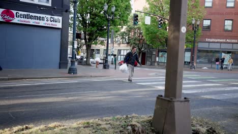 A-homeless-man-in-Portland,-Oregon,-crosses-Burnside-Street,-navigating-through-the-bustling-urban-environment,-carrying-his-belongings,-as-cars-and-pedestrians-move-around-him