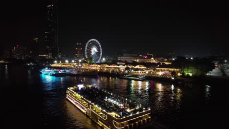 AERIAL:-Sightseeing-ferry-in-front-of-the-Asiatique-riverfront,-night-in-Bangkok