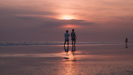 Two-friends-walk-on-beach-with-reflection-below-at-sunset-as-waves-crash-on-shore-with-wet-sand,-slow-motion-low-angle