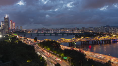 El-Tiempo-Del-Horizonte-Nocturno-De-Seúl-Transcurre-Con-Tráfico-Rápido-En-La-Autopista-Olympic-Daero,-Los-Puentes-Ferroviarios-Del-Río-Han-Y-Hangang-Y-El-Distrito-De-Negocios-De-Yeouido-Con-Imponentes-Rascacielos-De-63-Edificios-Y-Un-Cielo-Dramático