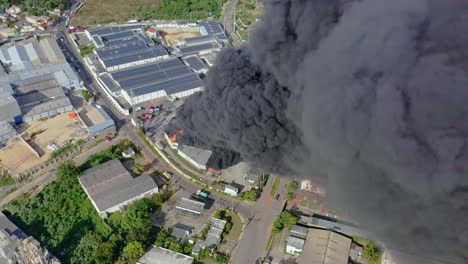 Aerial-drone-footage-of-a-factory-fire-in-the-Caribbean,-with-a-zoom-out-view-showing-thick-smoke-and-intense-flames-consuming-the-building,-capturing-the-dramatic-and-perilous-scene