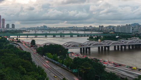 Day-to-Night-Timelapse-of-Cloudy-Seoul-Skyline-at-Sunset,-Busy-Car-Traffic-on-Olympic-daero-Highway,-View-of-Han-River-and-Hangang-Railway-Bridges-and-Yeouido-Skyscrapers-in-City-Downtown--High-Angle