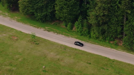 Coche-Circulando-Por-Una-Carretera-Asfaltada-Cerca-De-Bosques-Y-Campos-Verdes,-Seguimiento-De-Toma-Aérea-De-Cerca