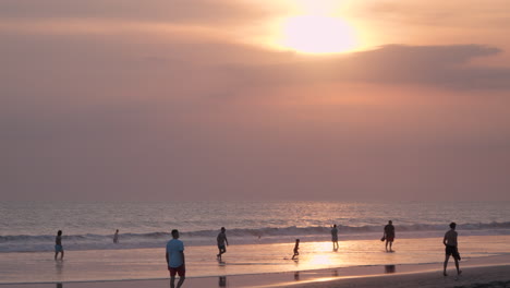 Families-play-and-run-in-shallow-water-flats-on-sandy-beach-at-sunset,-overcast-skies-diffuse-light-from-sun