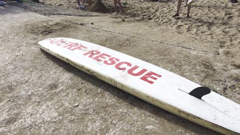 Ein-Surfbrett-Mit-Der-Aufschrift-„Surf-Rescue“-Liegt-Am-Strand-Und-Menschen-Laufen-Vorbei