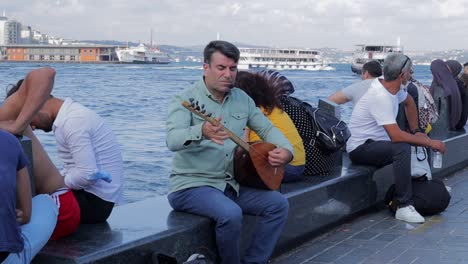 A-street-musician-and-others-sit-near-the-water-at-the-bay-of-Bosporus,-Istanbul,-capturing-a-vibrant-scene-of-local-culture-and-relaxation