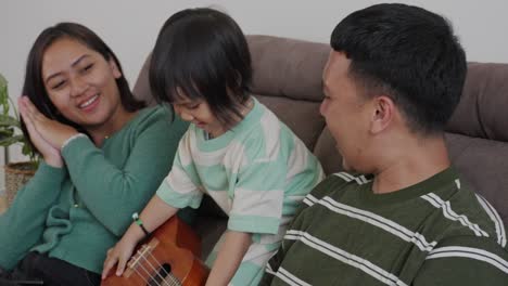 Little-Daughter-Playing-Guitar-Ukulele-And-Singing-With-Her-Parent-While-Sitting-Relaxed-At-Home