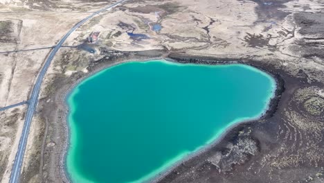 Drone-footage-captures-the-serene-beauty-of-a-teal-lake-in-Iceland,-nestled-within-a-natural-amphitheater-of-brown-rocky-terrain-and-green-grass