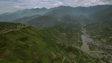 Vista-Aérea-De-Un-Paisaje-Montañoso-En-El-Norte-De-Vietnam-Con-Un-Pequeño-Pueblo-Y-Campos-De-Arroz-En-Terrazas-Junto-Al-Río