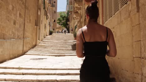 Vista-Posterior-De-Una-Mujer-Caminando-Por-Las-Escaleras-De-Un-Callejón-En-Un-Día-Caluroso-Y-Soleado-En-La-Valeta,-Malta.