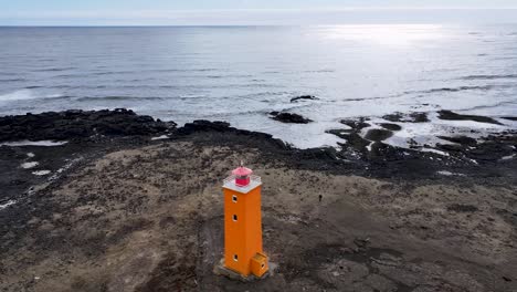 Das-Video-Zeigt-Eine-Atemberaubende-Nahaufnahme-Eines-Leuchtturms-An-Der-Küste-Islands,-Umgeben-Von-Schwarzem-Sand,-Felsvorsprüngen-Und-Majestätischen-Bergen-In-Der-Ferne.