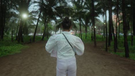 Woman-walking-through-a-park-pathway-surrounded-by-trees-in-the-early-evening