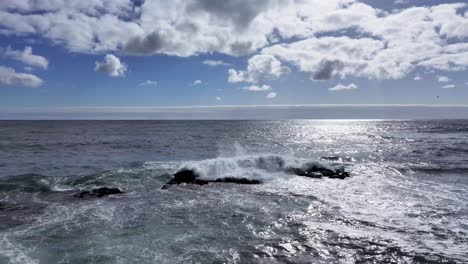 Tiefblaue-Meereswellen-Brechen-Gegen-Dunkle,-Schroffe-Felsen,-Während-Das-Sonnenlicht-Herabscheint-Und-Den-Dramatischen-Kontrast-Von-Farben-Und-Texturen-In-Diesem-Naturwunder-Hervorhebt