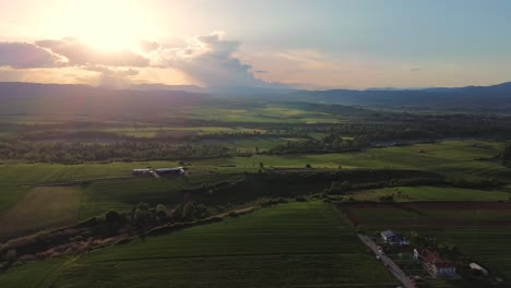 Green-rural-field,-aerial-sunset-between-clouds-skyline,-modern-village,-Greece