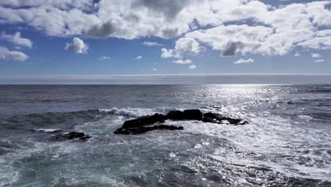 El-Video-Captura-El-Impresionante-Poder-De-Las-Olas-Cuando-Se-Encuentran-Con-La-Costa-Rocosa,-Rodeadas-Por-Un-Cielo-Azul-Claro-Sereno-Y-La-Luz-Del-Sol-Brillando-En-La-Superficie-Del-Agua.