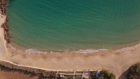 Drone-top-down-rising-from-pristine-golden-sand-beach-with-clear-turquoise-blue-ocean-water-waves-crashing-on-shore-in-Donousa-Greece