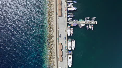 Barcos-Atracados-En-Un-Muelle-En-La-Costa-De-Cerdeña,-Italia,-Con-Agua-Azul-Clara,-Vista-Aérea