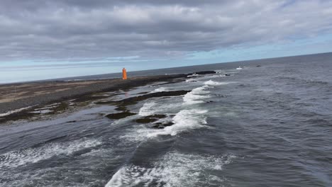 The-aerial-footage-features-a-stunning-lighthouse-on-Iceland's-coast,-surrounded-by-a-unique-landscape-of-black-sand,-rocks,-and-ocean,-with-majestic-mountains-in-the-distance