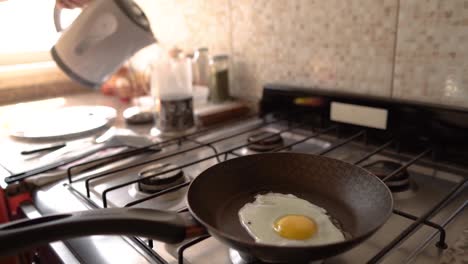 Close-up-of-a-fried-egg-being-cooked-in-a-pan-on-a-gas-stove-and-hot-tea-being-served-on-a-jug