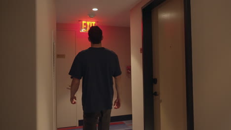 A-young-man-follows-a-big-red-neon-exit-sign-through-a-building-corridor,-trying-to-find-his-way-out