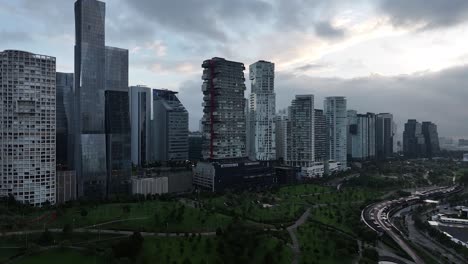 Panoramic-aerial-of-Parque-La-Mexicana-at-dusk,-pan-left-revealing-the-dynamic-contrast-between-nature-and-the-city