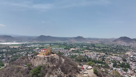 Ascending-drone-aerial-of-San-Miguel-Hill-Chapel-in-Atlixco,-Puebla-reveals-valley-cityscape-and-mountain-range-on-horizon