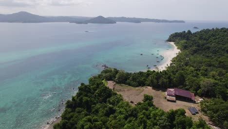 Aerial-panoramic-view-of-a-serene-tropical-coastline-of-Coiba-Island-with-clear-blue-waters,-lush-greenery,-and-a-small-building-nestled-near-the-shore-under-a-blue-sky