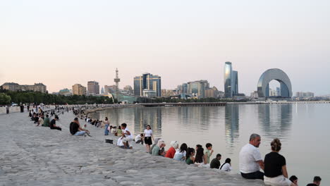 La-Gente-Se-Sienta-En-El-Paseo-Marítimo-Junto-Al-Agua-En-Bakú,-Con-Un-Horizonte-De-Edificios-Modernos