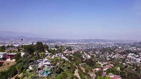 Impresionante-Vista-Aérea-De-Casas-En-La-Ladera-De-Una-Colina-Con-Exuberante-Vegetación,-Con-Vistas-Al-Extenso-Paisaje-Urbano-De-Los-Ángeles-Bajo-Un-Cielo-Azul-Claro