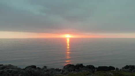 Tidal-pool-near-rocky-beach-of-Farstadsanden-beach-at-sunset-in-Norway,-drone-dolly-to-last-light-glow-below-clouds