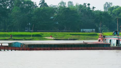 A-loaded-cargo-ship-in-strong-tide-at-a-flooded-river-in-rural-Bangladesh,-amidst-lush-green-surroundings