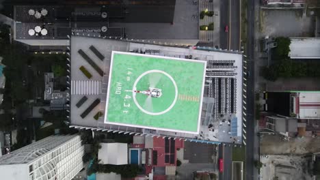 Top-down-aerial-view-of-a-helicopter-with-rotor-spinning-on-a-symmetrical-helipad-at-the-top-of-a-skyscraper-surrounded-by-tall-buildings-of-the-city