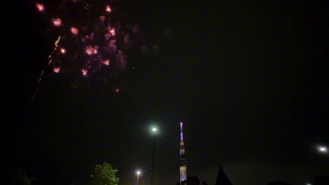 Ein-Feuerwerk-In-Leuchtenden-Farben-Erstrahlt-Nachts-über-Der-Skyline-Einer-Stadt-Mit-Einem-Hohen-Beleuchteten-Turm-Im-Blick