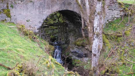 Idyllic-stream-of-water-flowing-under-old-stone-bridge-arch-in-rural-countryside-of-Scotland-UK