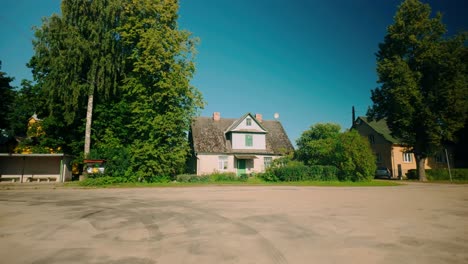 A-large,-old-Baltic-house-stands-next-to-concrete-road,-surrounded-by-lush-green-trees-during-summer