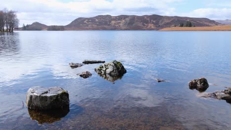 Beautiful-loch-with-crystal-clear-freshwater,-rocky-boulders-and-mountains-in-the-distance-in-highlands-of-Scotland-UK