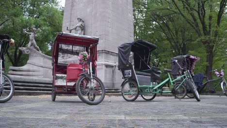 Estacionamiento-De-Rickshaw-Tuk-Tuk-En-El-Parque-Central-De-Nueva-York-Detrás-De-Un-Monumento-Con-Un-árbol-De-Fondo