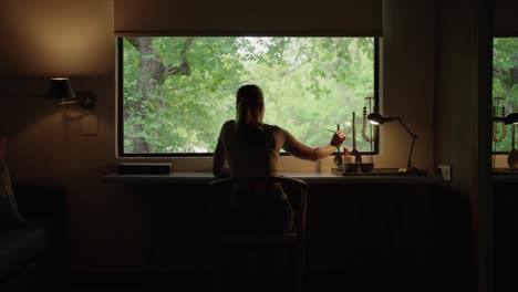 A-woman-picking-up-a-pencil-and-writing-on-a-notepad-at-a-desk-in-front-of-a-wide-window-with-a-natural-green-landscape-outside