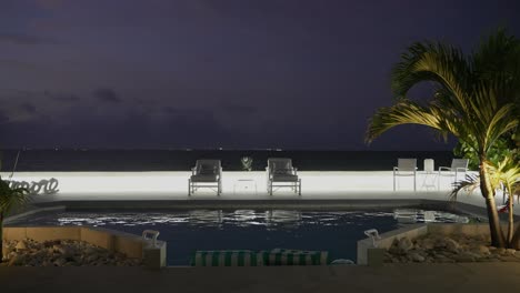 Toma-De-Una-Elegante-Piscina-De-Lujo-Junto-A-La-Playa-Durante-La-Noche-Con-Palmeras