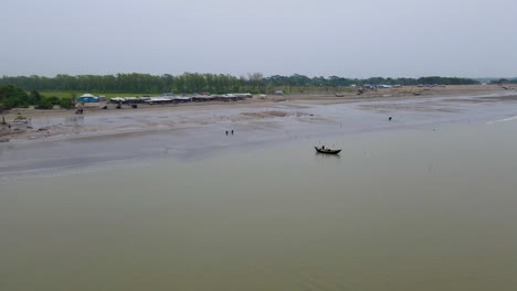 Luftflug-über-Den-Strand-Von-Kuakata-Mit-Einem-Kleinen-Hölzernen-Fischerboot-An-Einem-Grauen-Tag,-Lebur-Char,-Kleiner-Markt-Im-Hintergrund,-Kopierraum