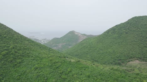 Drone-flying-through-mountains-filled-with-greenery-of-Saint-Martin-during-cloudy-day-in-Carribean-Island