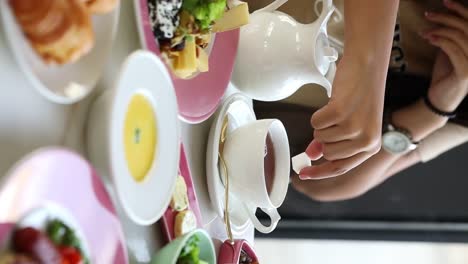 A-woman-puts-a-cube-of-sugar-in-her-tea-beside-a-delicious-breakfast-in-fancy-restaurant-close-up-shot,-vertical-video,-insert-shot